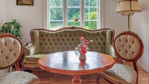 Een kamer met een groene gestoffeerde bank, een ronde houten tafel met een bloevas, en twee stoelen met bekleden zittingen.