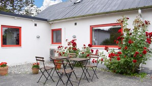 En terrasse med et bord og stole, en hvid bygning med rødlakerede vinduer, og en rosenbusk.