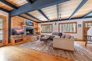 A cozy cabin interior with a fireplace, a sofa, a television, and large windows.