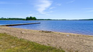 A lakeside view with a wooden dock extending into the water, surrounded by a sandy beach and sparse grass.