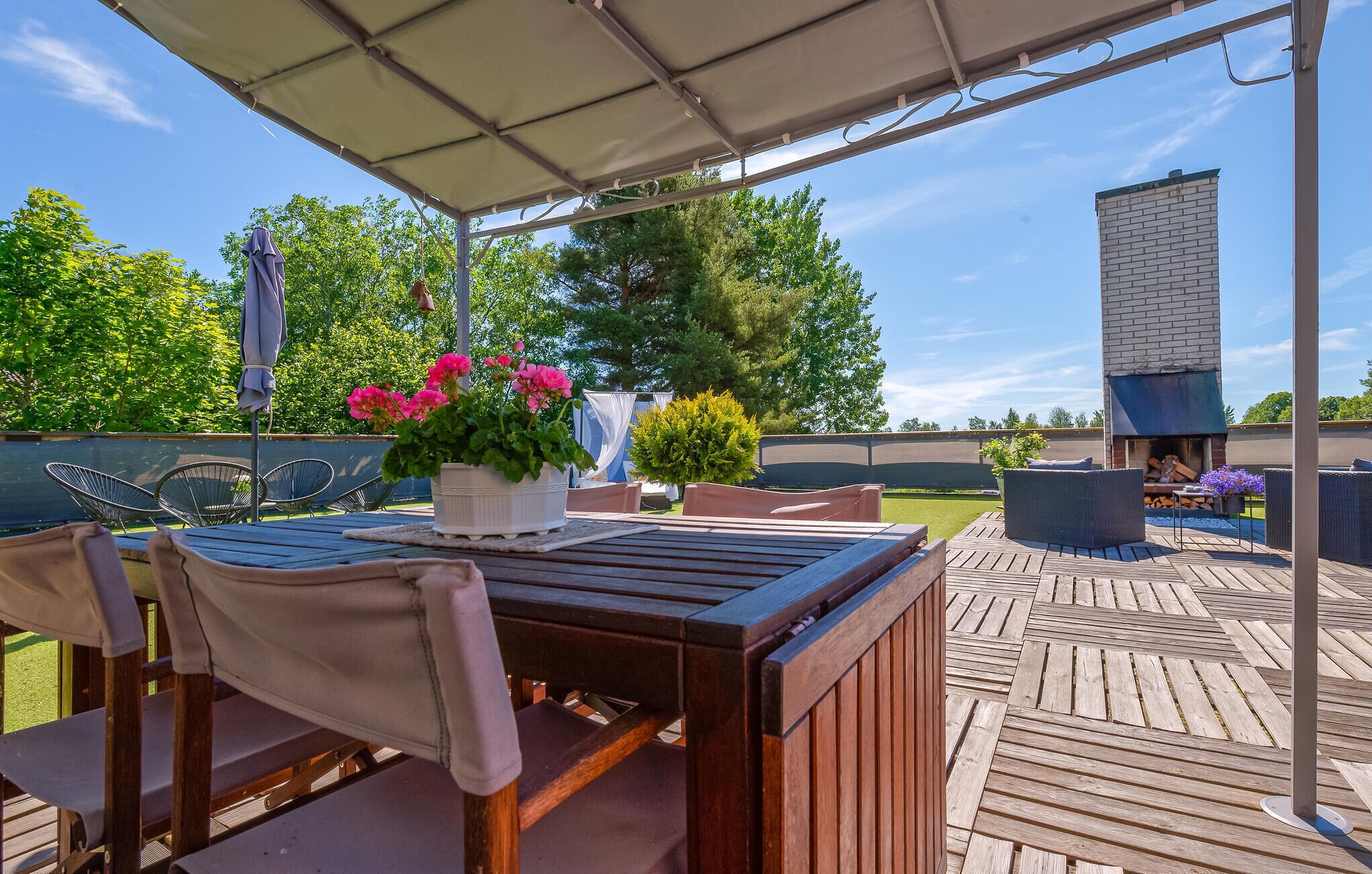 A covered outdoor patio with a wooden table and chairs, surrounded by potted plants and a barbecue area.