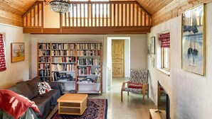 Uma sala de estar acolhedora com teto de madeira alto, lareira, estante cheia de livros, sofá confortável e tapete estampado.
