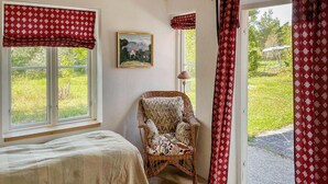 Quarto com cama, cadeira de vime, um quadro e vista para um jardim.