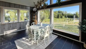 Un comedor con una mesa dispuesta para cuatro personas, ventanas amplias que ofrecen vistas a un paisaje cubierto de hierba, y un techo decorado con cuernos de ciervo.