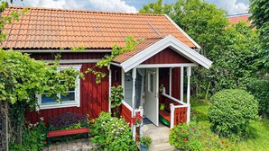 Une maison en bois rouge, avec un toit en tuiles, un porche et un jardin où poussent différentes plantes et fleurs.