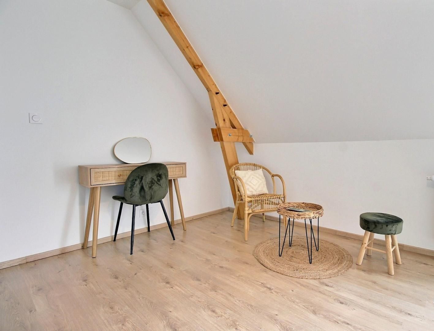 Quarto estilo loft com teto inclinado, escrivaninha de madeira com espelho, cadeira preta, cadeira de vime, mesa redonda pequena e radiador.