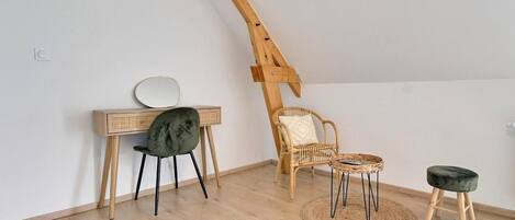 Quarto estilo loft com teto inclinado, escrivaninha de madeira com espelho, cadeira preta, cadeira de vime, mesa redonda pequena e radiador.