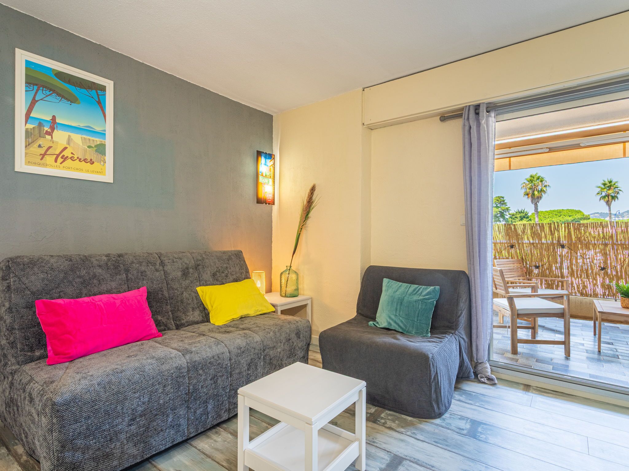 A modern living room with a grey sofa, a small white table, and a grey armchair. There is a framed picture on the wall and a window with a view of palm trees and a beach.