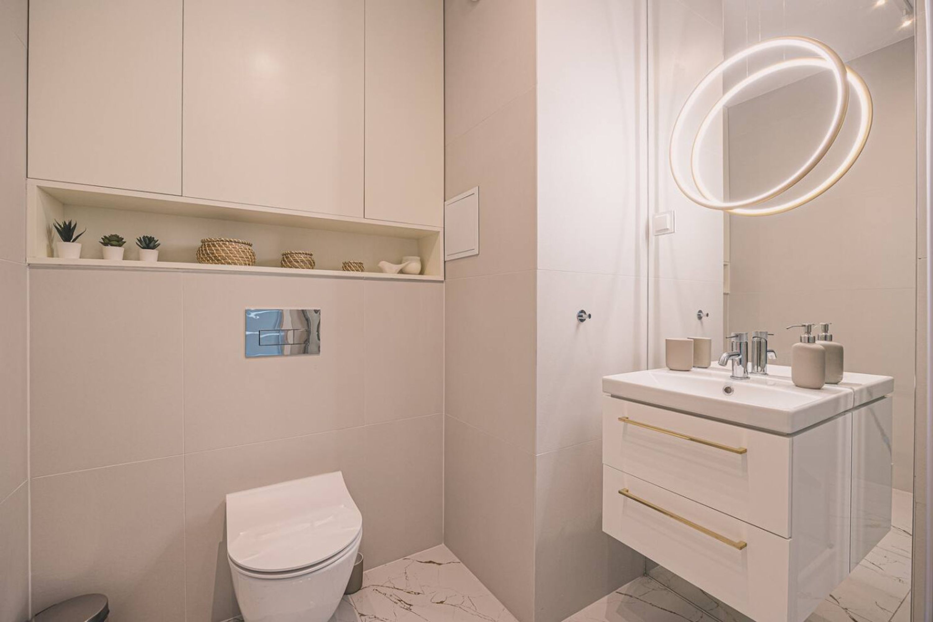 A modern bathroom with a toilet, a vanity with a mirror and lights, and shelves with plants and toiletries.