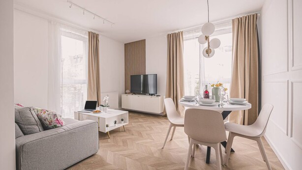 A modern living room with a sofa, a dining table with chairs, a TV, and a white cabinet. - Blanco Apartament Gara Balkon (Bydgoszcz)