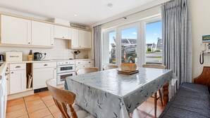 Eine moderne Küche mit weißen Schränken, einem Tisch mit Tischdecke und Blick auf die Straße durch große Fenster.