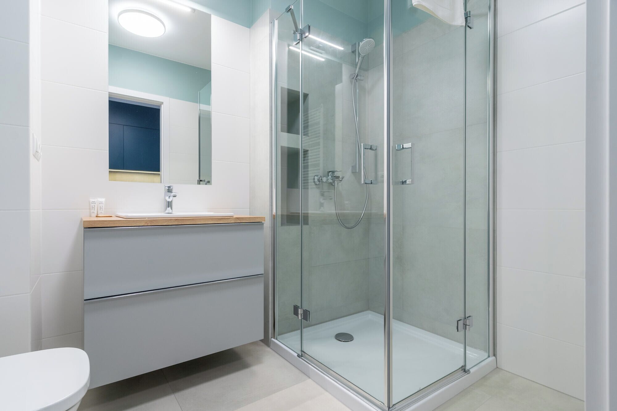 Ein modernes Badezimmer mit einer Glaskabine zur Dusche, einem weißen Waschbecken mit Holzablage und einem Spiegel darüber.