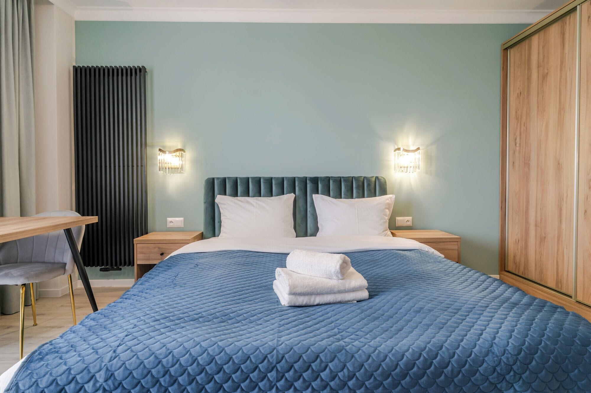 Ein ordentlich bezogenes Bett mit einer blauen Steppdecke, zwei weißen Kissen und einem gefalteten Handtuch. Zwei Nachttische mit Lampen, ein Holztisch und ein Kleiderschrank sind ebenfalls vorhanden.