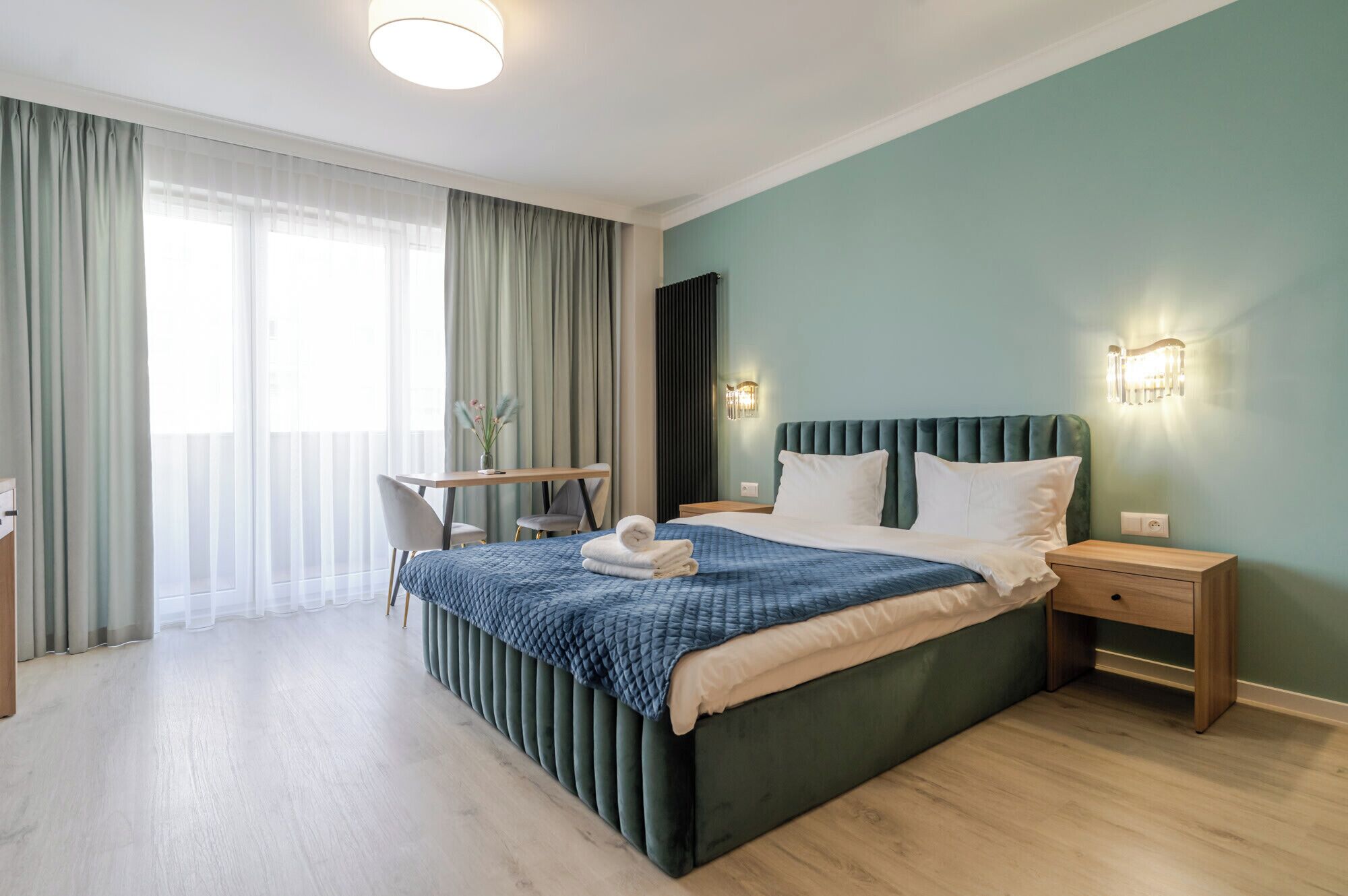 Ein modernes Hotelzimmer mit einem großen Bett, einem Schreibtisch und einem Stuhl. Es gibt ein Fenster mit Vorhängen, eine Wandleuchte und eine Heizung.