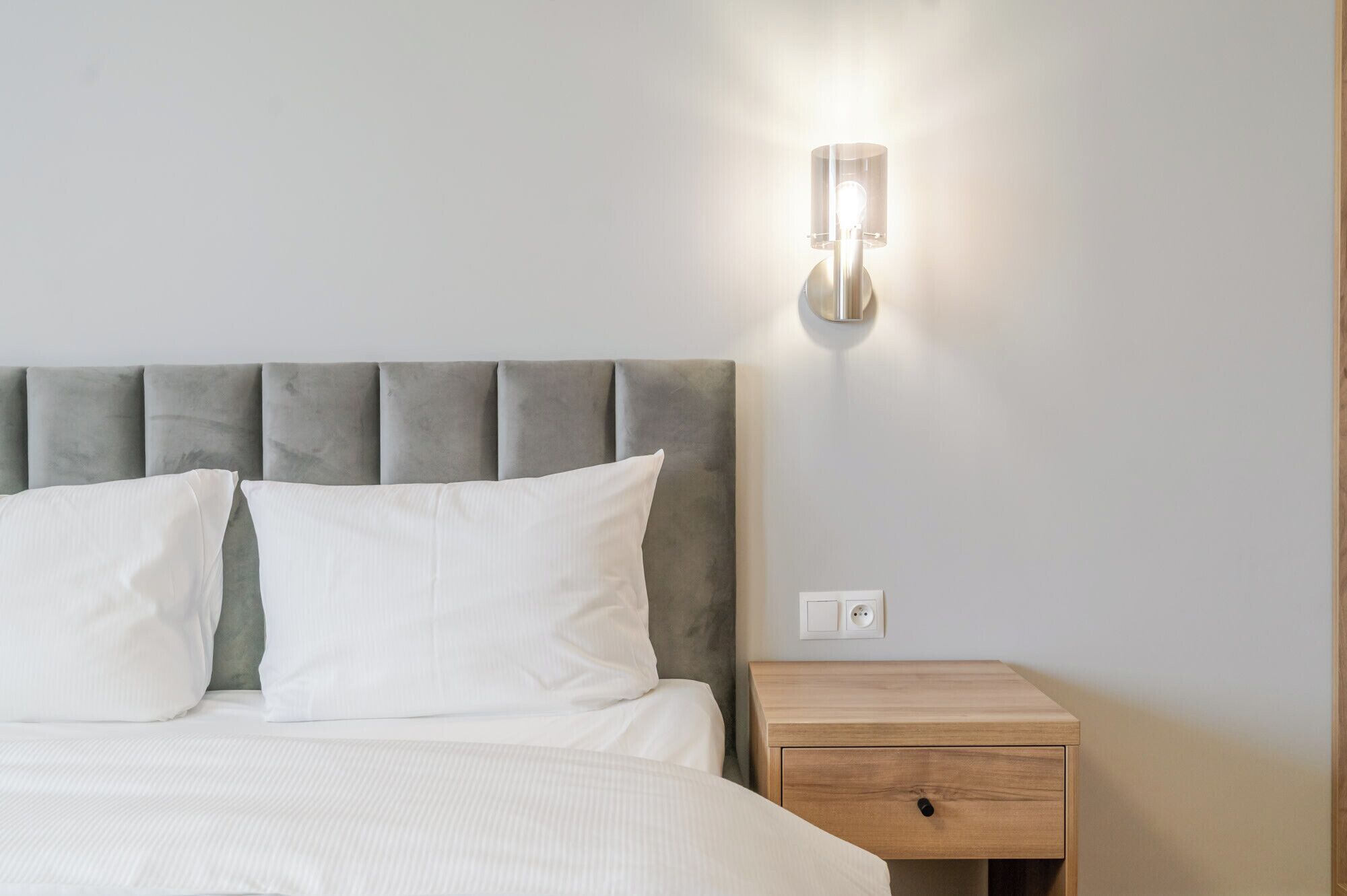 Una cama con cabecera tapizada en gris, ropa de cama blanca y una mesita de noche con cajón.