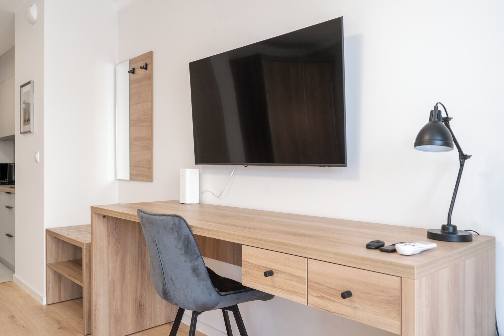 Een moderne kantoorruimte met een houten bureau, een grijze stoel, een wandgemonteerde televisie, een bureaulamp en een klein wit apparaat op het bureau.