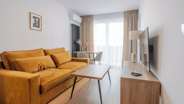 A modern hotel room with a mustard-colored sofa, a wooden coffee table, a small desk with a lamp, a television, and a view of the outdoors through large windows. - Business Deluxe Studio 19 (Katowice)