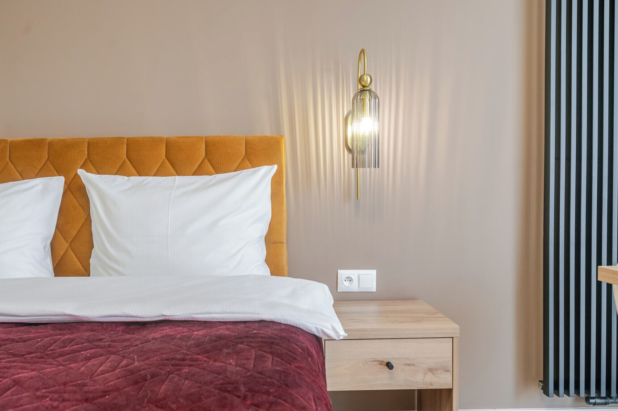 A neatly made bed with a red blanket, a wooden nightstand with a drawer, and a wall-mounted light fixture.