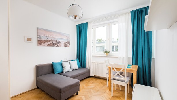 A modern living room with a grey sofa, teal curtains, a wooden herringbone floor, a white radiator, and a window with a view of buildings.