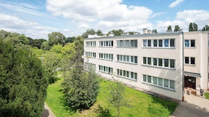 A modern multi-story building with a flat roof, surrounded by lush greenery and trees.