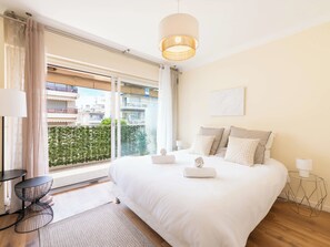 A hotel room with a large bed, a modern lamp, a small side table, and a view of the city through large windows. - 1-Br with Terrace – Palm Beach, Close to Beaches (Cannes)