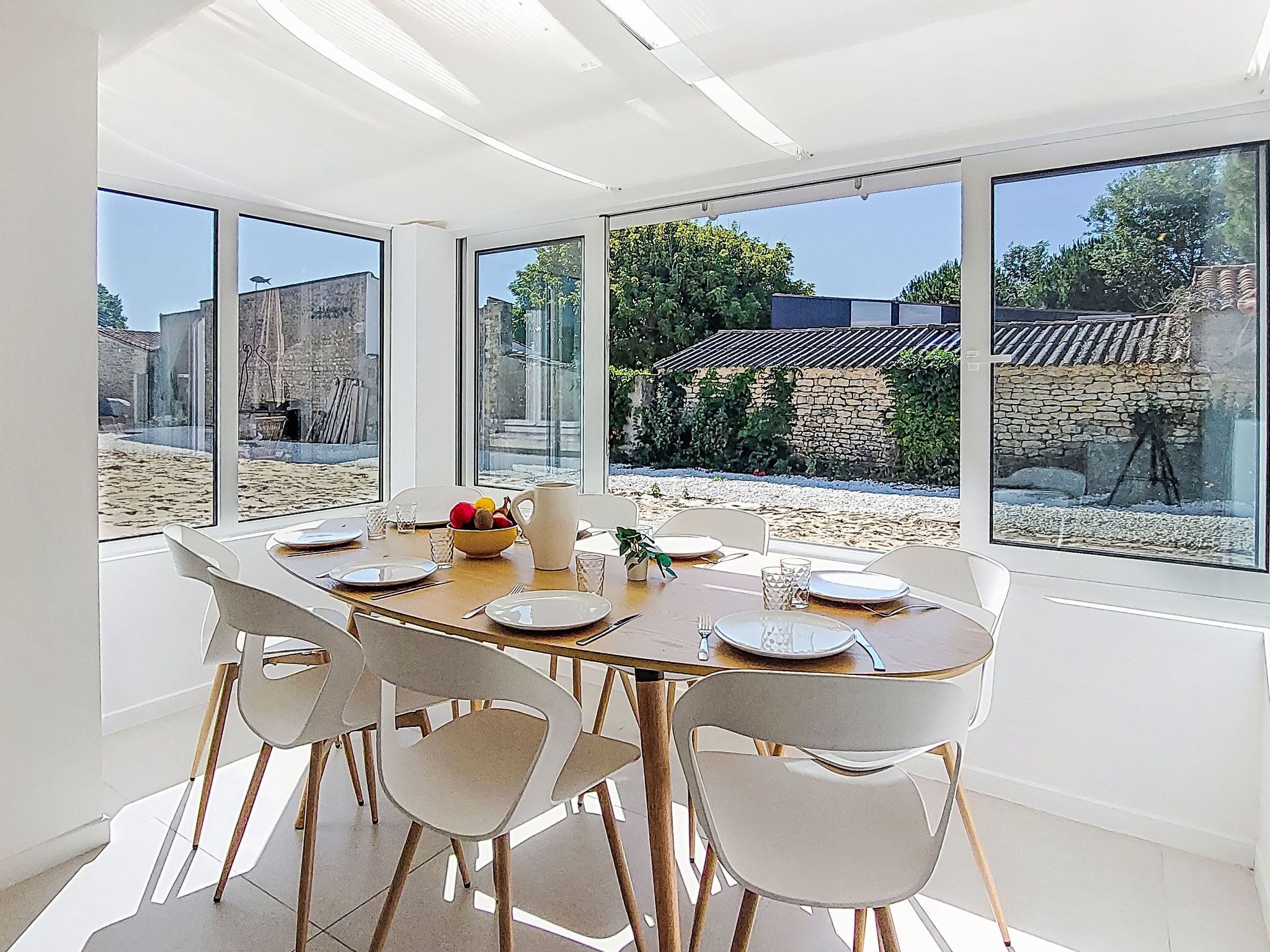 Ein Essbereich mit einem großen Tisch, gedeckt für sechs Personen, umgeben von weißen Stühlen mit Holzbeinen. Große Fenster bieten Blick auf einen Garten und umliegende Gebäude.