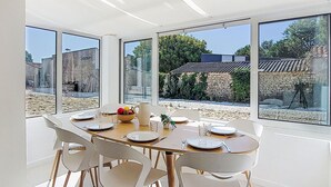Un espace repas avec une grande table dressée pour six personnes, entourée de chaises blanches aux pieds en bois. De grandes fenêtres offrent une vue sur un jardin et les bâtiments environnants.