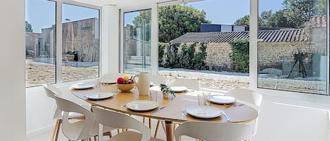 A dining area with a large table set for six, surrounded by white chairs with wooden legs. Large windows offer views of a garden and buildings outside.