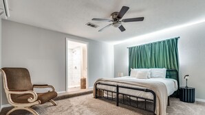 Un dormitorio con cama, una silla, ventilador de techo y una ventana con cortinas.