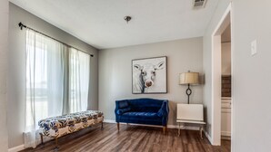 Una habitación con un sofá de terciopelo azul, un banco con estampado floral, un cuadro de una vaca y una lámpara sobre una mesita.