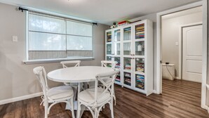Una habitación con una mesa redonda blanca y cuatro sillas blancas, un armario blanco con puertas de cristal y una ventana con persianas.