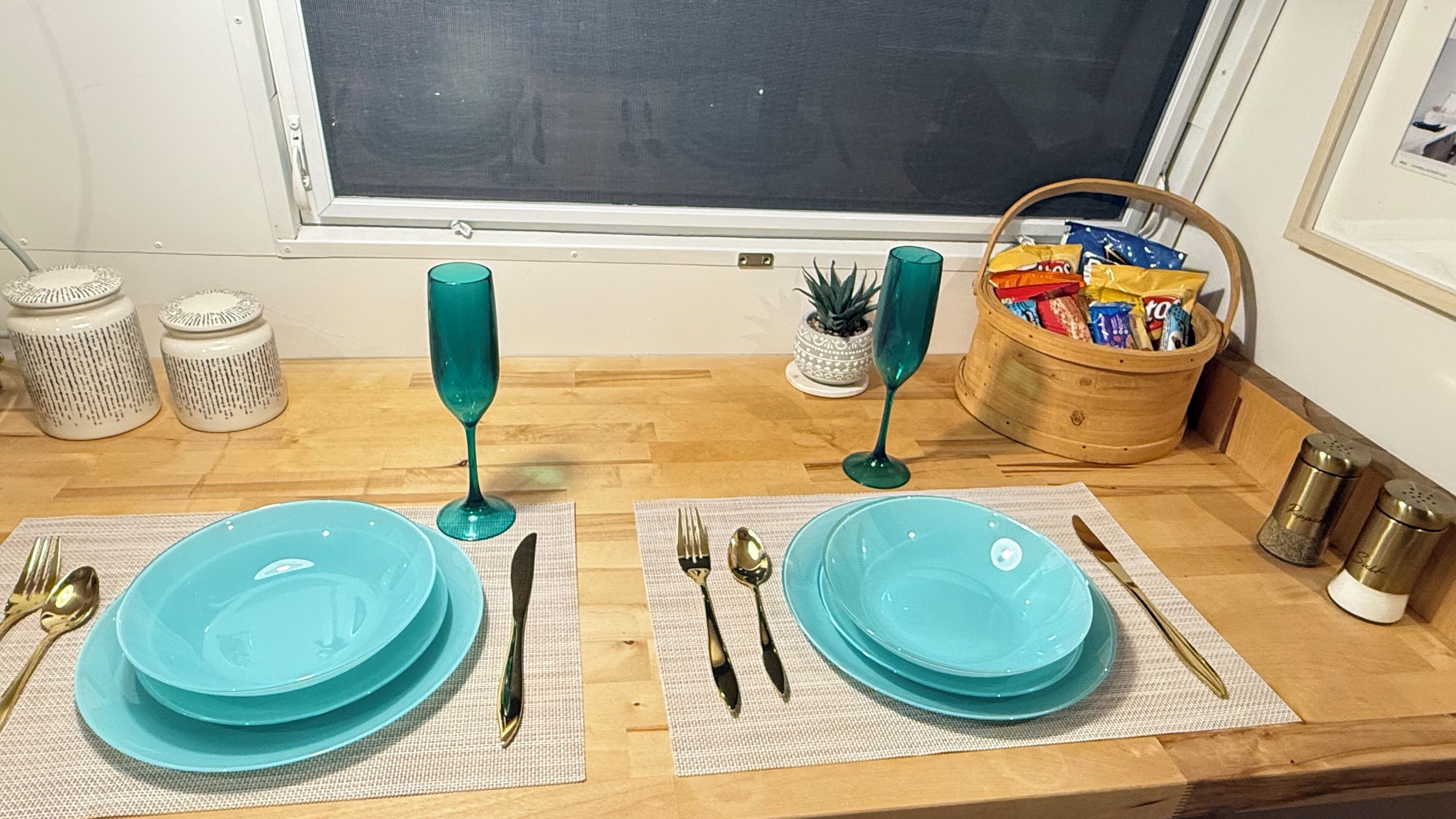 Zona de menjador amb dos servis de plats turquesa, coberts i vasos posats sobre proteccions de tafet. Es veu una cistella de fusta amb aperitius i una petita planta en test a l'esfondri.