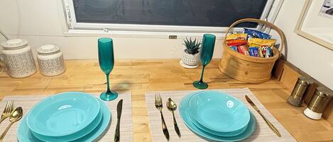 Et spiseområde med to sæt turkise tallerkener, bestik og glas på vævede bordskåner. En trækurv med snacks og en lille potteplante ses i baggrunden.