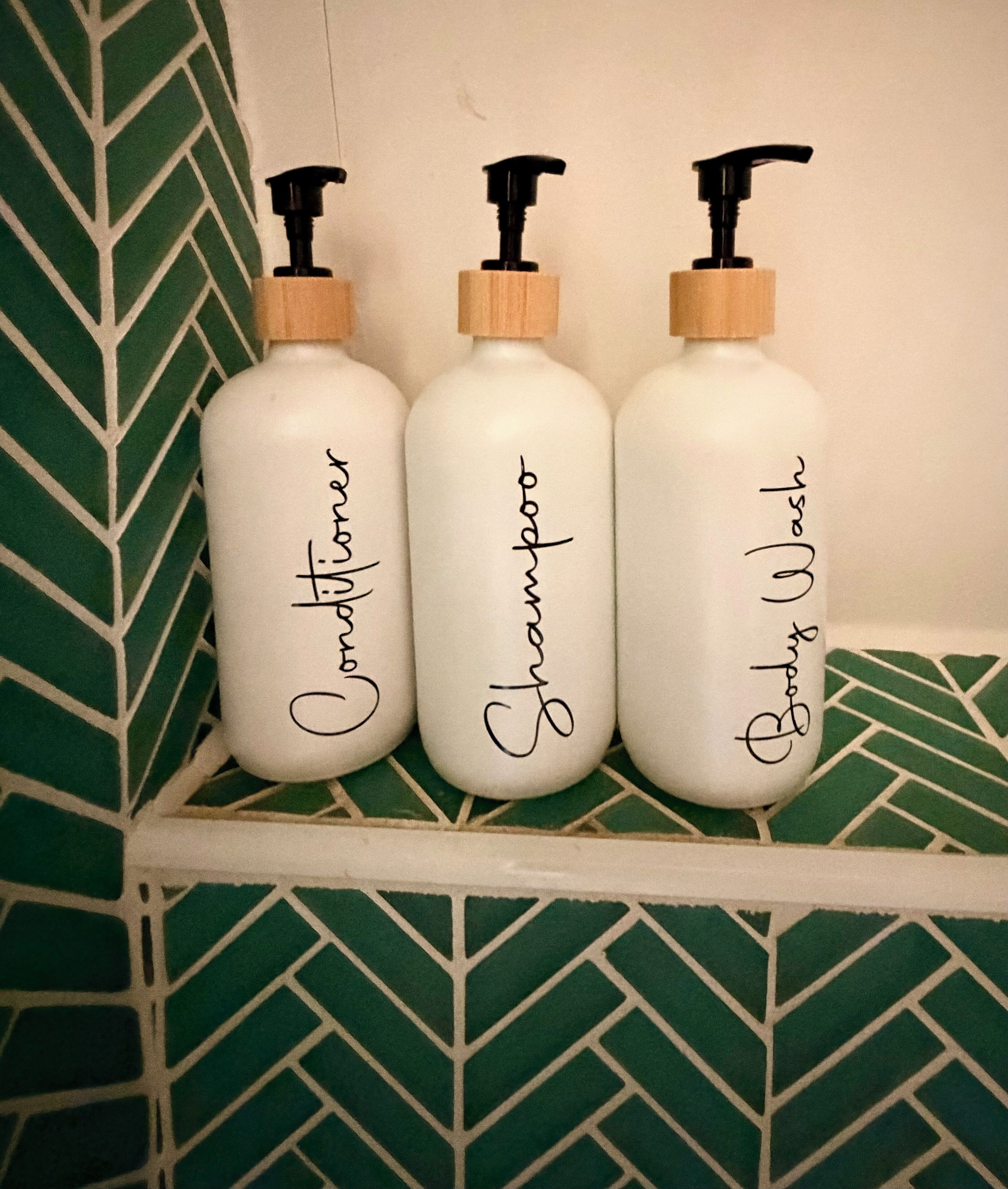 Tres botellas blancas con bombas negras y etiquetas, cada una con un tipo diferente de champú, colocadas sobre una estantería frente a una pared verde con diseño.