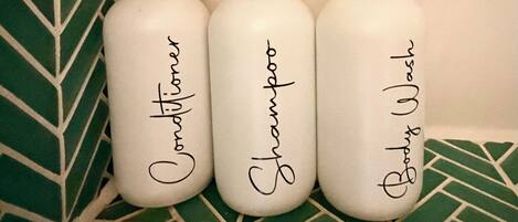 Tres botellas blancas con bombas negras y etiquetas, cada una con un tipo diferente de champú, colocadas sobre una estantería frente a una pared verde con diseño.