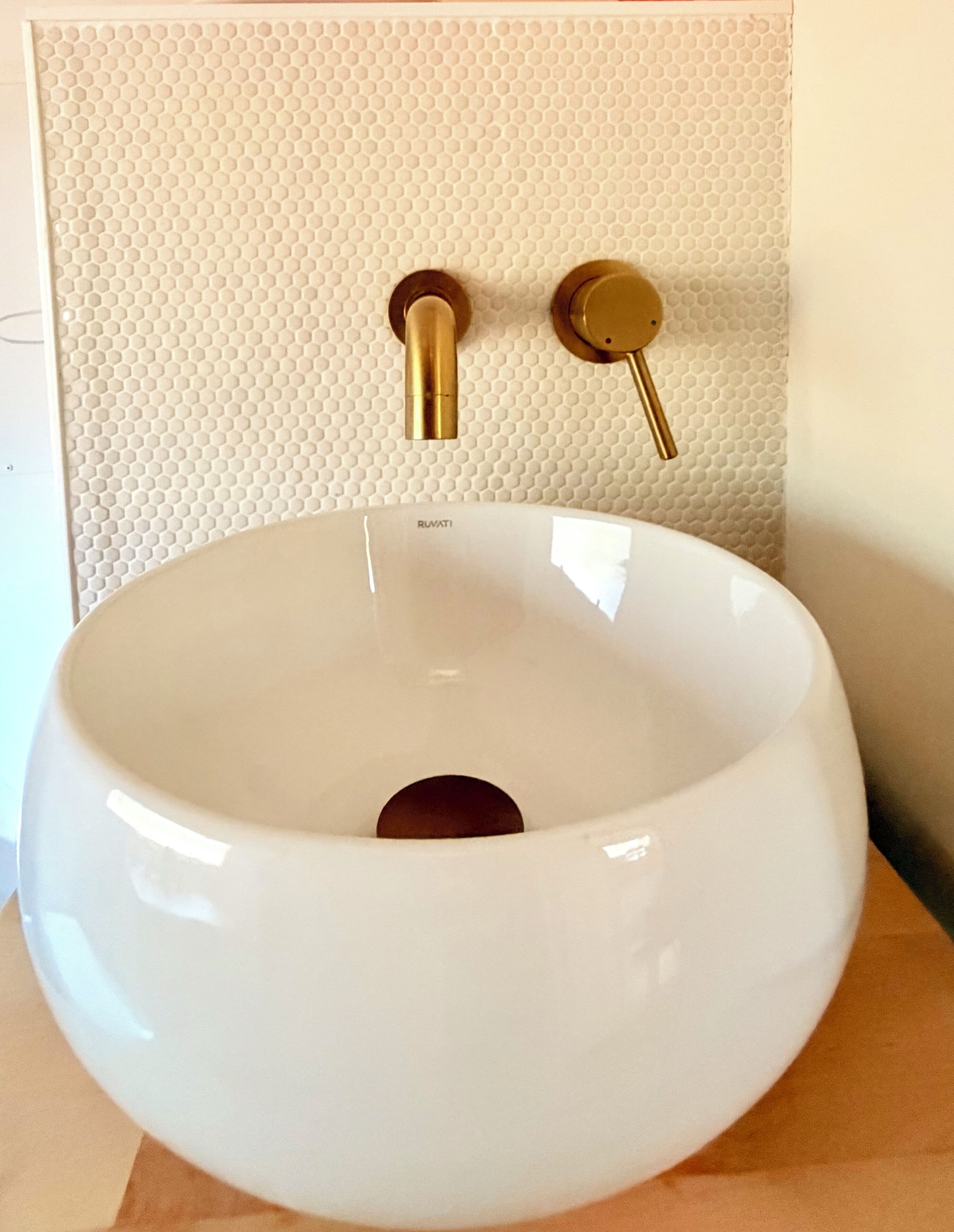 A white ceramic sink with a gold faucet and a gold handle, mounted on a textured wall.