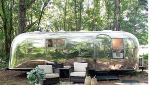 A silver RV with a reflective exterior, situated in a wooded area. It features a small window, a door, and is equipped with outdoor furniture including a table and chairs.