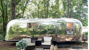A silver RV with a small window, surrounded by wicker furniture and a potted plant.