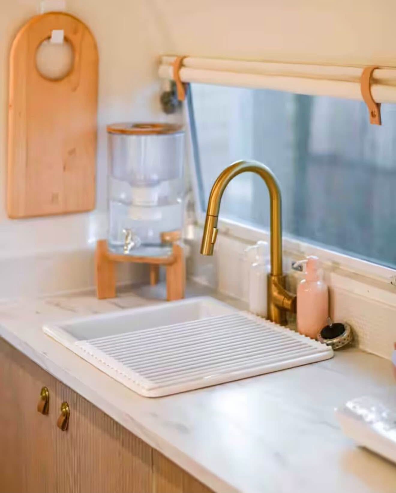 A kitchen with a white countertop, a gold faucet, a wooden cutting board, a water dispenser, and a window.