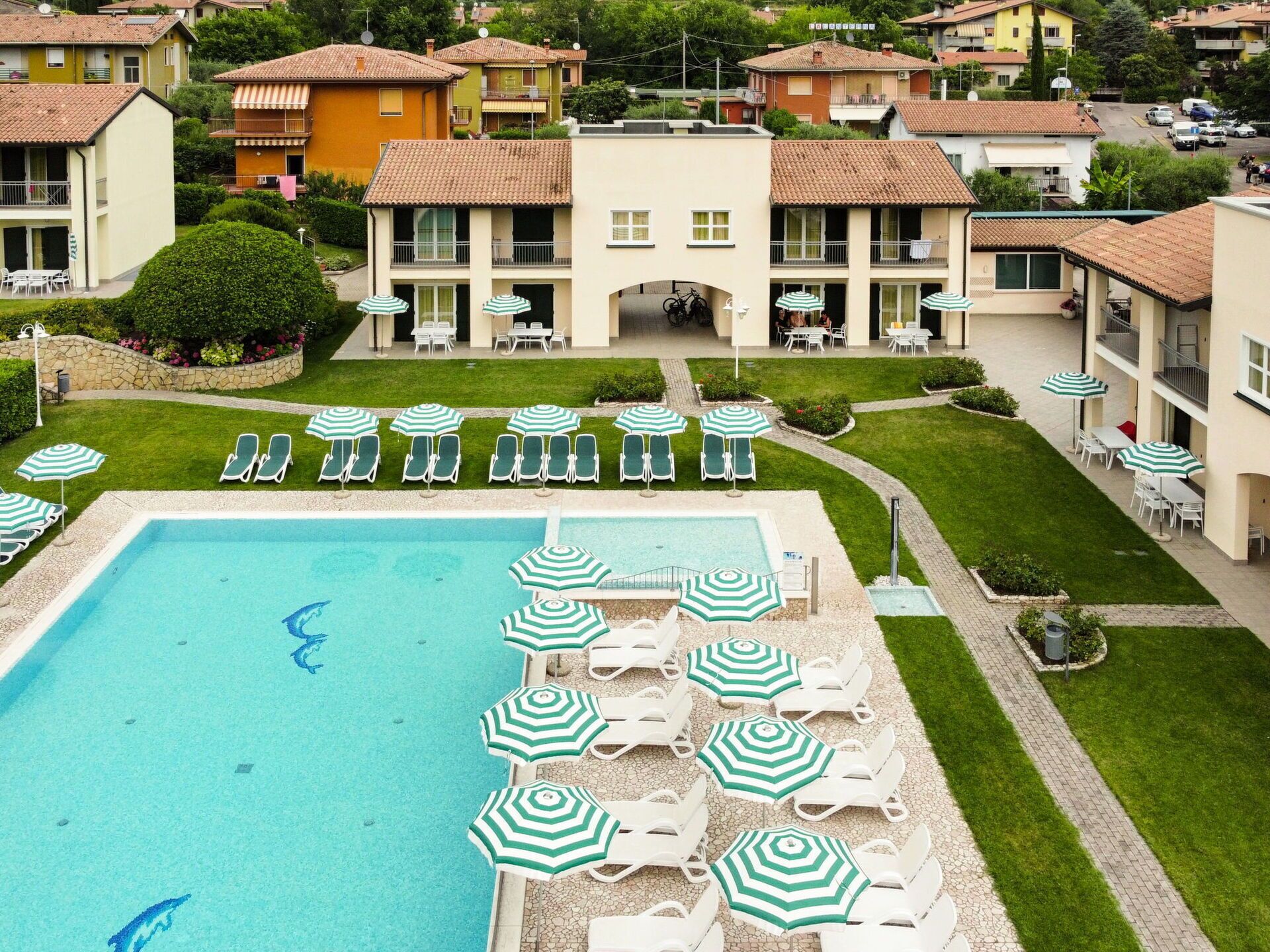 Une piscine entourée de chaises longues et de parasols, avec une pelouse bien entretenue et des immeubles d’habitation en arrière-plan.