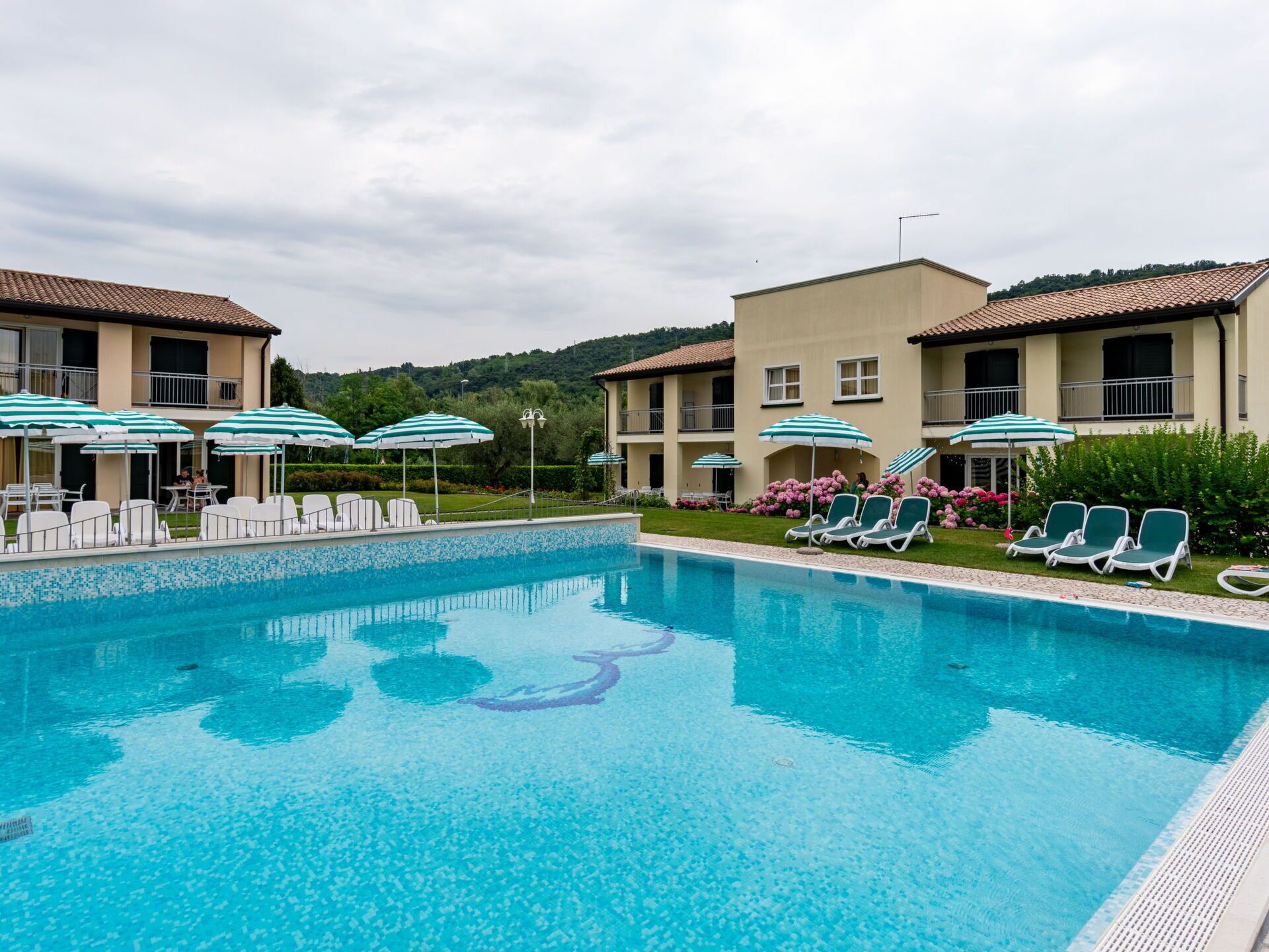 A swimming pool with lounge chairs and umbrellas, surrounded by buildings with balconies and a hilly landscape in the background.