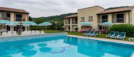 A swimming pool with lounge chairs and umbrellas, surrounded by buildings with balconies and a hilly landscape in the background.