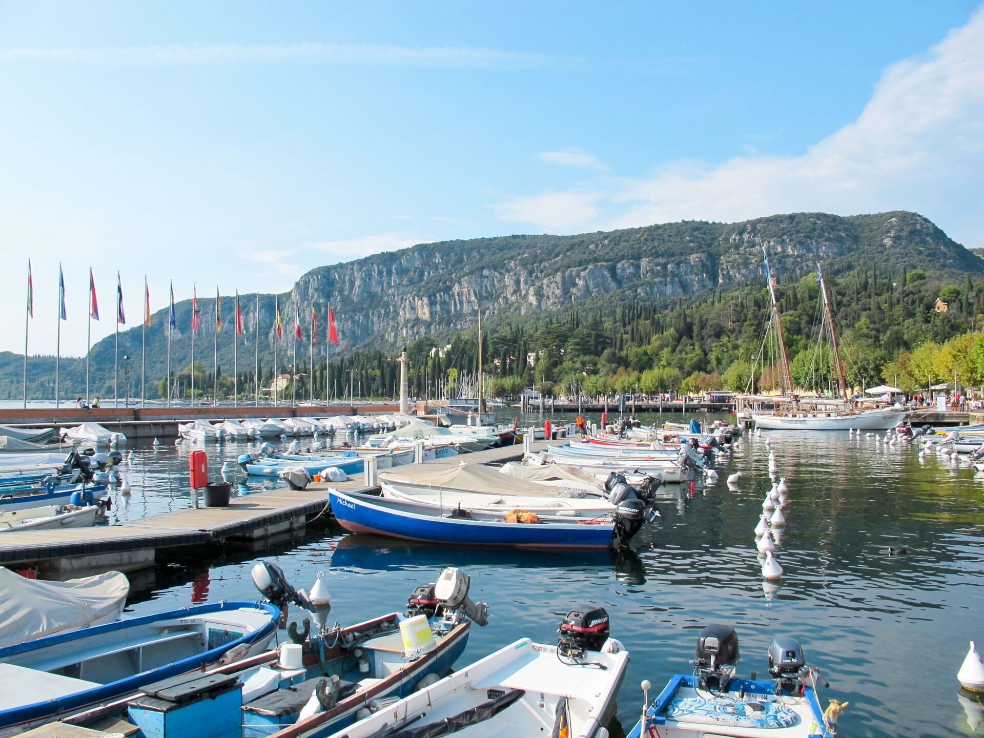 A marina with various boats docked, a mountainous backdrop, and flags on poles.