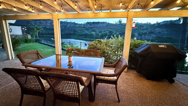 An outdoor patio with a glass table, wicker chairs, a grill, and a view of a lake and hills.