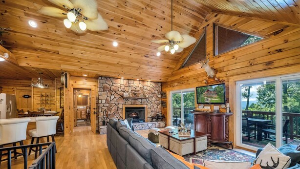 A spacious living room with a stone fireplace, wooden ceiling, and a large TV. - Wraparound Views and Forest Feels with Game Room, Pool Table and Hot Tub! (Grassy Creek)