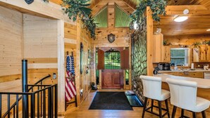A wooden cabin interior with a kitchen area, a dining table with chairs, and a staircase leading to an upper level. - Wraparound Views and Forest Feels with Game Room, Pool Table and Hot Tub! (Grassy Creek)