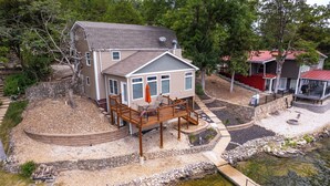 A two-story house with a deck, a stone retaining wall, and a small pond. - Charming Lake of the Ozarks Cabin, 3BR + Loft, Dock & Fun for All! Sleeps 10 (Rocky Mount)