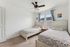 A bedroom with two single beds, a ceiling fan, a window with curtains, and a small table. - Spacious 4 BR Condo with a Garage & Private Decks (Wildwood)
