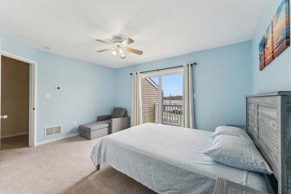 A bedroom with a bed, a ceiling fan, a window with curtains, a door, and a small seating area. - Spacious 4 BR Condo with a Garage & Private Decks (Wildwood)
