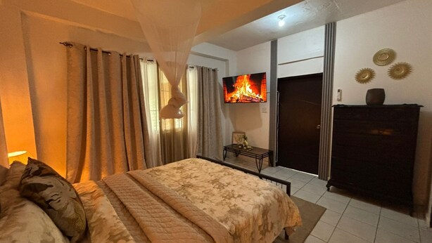 Une chambre à coucher avec un lit, des rideaux, une télévision, une commode et une porte.
